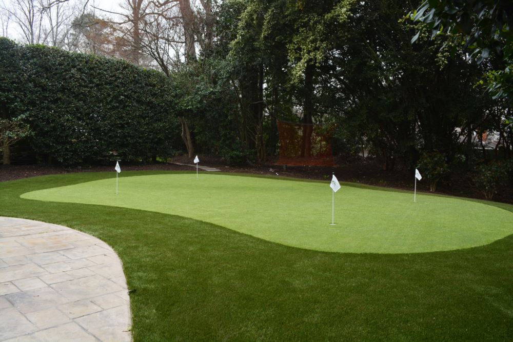 Custom artificial turf putting green with multiple golf practice holes, installed in a landscaped backyard next to a curved stone patio and surrounded by trees.